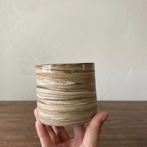 May include: A cylindrical ceramic container with a wood grain pattern in shades of brown and tan. The handmade pottery has a smooth, glossy finish and is held by a hand. The container is suitable for holding small plants or as a decorative item.