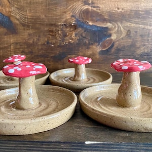 May include: Four ceramic ring dishes shaped like mushrooms with red and white speckled tops. The dishes are a speckled beige color.
