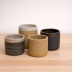 May include: Four handcrafted ceramic tumblers in varying sizes and colors. The tumblers are cylindrical with a speckled tan, black, and cream finish. They are arranged on a light wood surface against a white background.