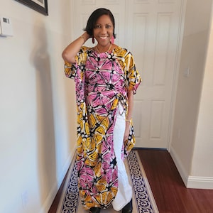 May include: A woman wearing a pink, yellow, and black patterned long-sleeved top with a white skirt. The top has a high neckline and a loose fit. The skirt is a high-waisted, flowy style.