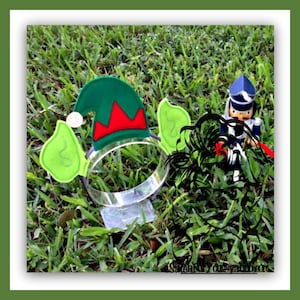 May include: A green felt elf hat with red trim and green felt ears attached to a clear plastic headband. The headband is sitting on a bed of green grass.
