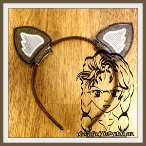 May include: A brown headband with two gray felt cat ears. The ears have a white felt inner lining. The headband is on a wooden surface.