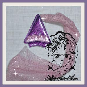Puede incluir: Una diadema de tela con purpurina morada y rosa con un adorno de purpurina morada en forma de triángulo. La diadema está decorada con un diseño de purpurina rosa y blanca. El texto "aStitchForYou@yahoo.com" está impreso en la tela.