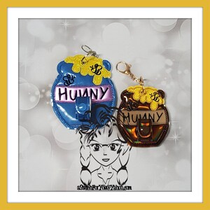 May include: Two honey pot-shaped keychains with the word "Hunny" on them. The keychains are made of fabric and have a shiny, iridescent finish. One keychain is blue and the other is brown.