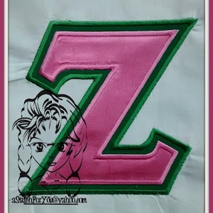 Könnte beinhalten: Ein pinkfarbener Buchstabe Z mit grünem Umriss. Der Buchstabe ist auf einen weißen Stoffhintergrund gestickt.