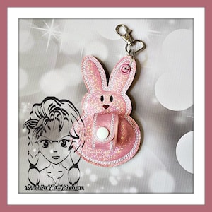 May include: Pink glitter bunny shaped keychain with a small pocket and a white button. The keychain has a silver ring and a silver clip.