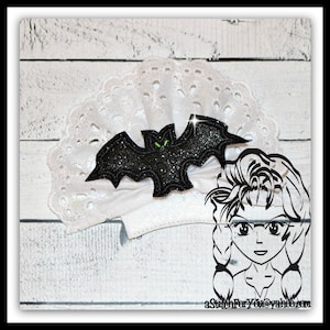 Peut inclure: Un bandeau en dentelle blanche orné d'une chauve-souris noire pailletée avec des yeux verts. La chauve-souris est centrée sur un nœud blanc. Le fond est une surface en bois blanc. Une illustration de dessin animé en noir et blanc est à droite.