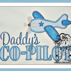 May include: Embroidered design featuring a blue airplane with a white star and a blue and white gingham pattern. The text "Daddy's Co-Pilot" is also embroidered in blue and white gingham.