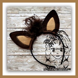 Puede incluir: Diadema de fieltro marrón con un par de orejas de fieltro marrón con el interior de fieltro beige. La diadema tiene una pluma marrón en la parte superior. La diadema está sobre un fondo de madera blanca.