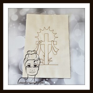Op de afbeelding: Een witte stof met een bruin geborduurd ontwerp van een kruis met een zonneschijn erachter. Het kruis wordt omringd door twee linten. De stof bevindt zich in een frame.