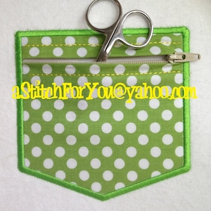 May include: A green fabric pocket with white polka dots and a zipper. The pocket has a green stitched border and a yellow stitched line around the zipper. A pair of small silver scissors is sitting on top of the zipper.