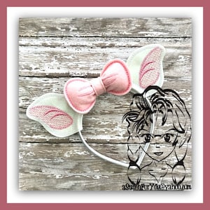 May include: A white headband with pink felt ears and a pink bow. The headband is on a wooden surface.