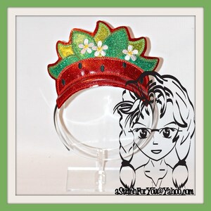 May include: A red and green headband with a strawberry design. The headband is decorated with glitter and has a white plastic stand.