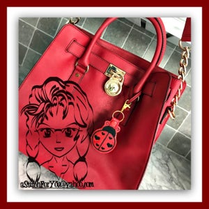 Può includere: Borsa a mano in pelle rossa con una tracolla a catena dorata e una serratura dorata. Un'immagine di cartone animato in bianco e nero di una ragazza con gli occhiali è sul davanti della borsa. Un portachiavi rosso a forma di coccinella è attaccato alla borsa.