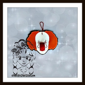 May include: A felt ornament shaped like a clown's face with orange hair, white skin, and a red nose. The clown's eyes are black and the mouth is open with a red tongue sticking out. The ornament has a loop at the top for hanging.