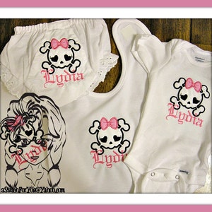 May include: A set of three white baby clothes with pink skull and crossbones designs and the name "Lydia" embroidered on each piece. The set includes a onesie, a bib, and a diaper cover.