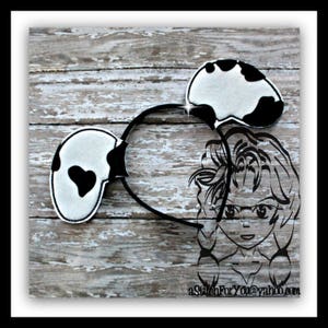 May include: Black and white cow-print headband with a heart-shaped design on one ear. The headband is on a wooden background.