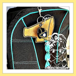 May include: A black backpack with a turquoise trim and a gold and black megaphone keychain attached to the zipper. The keychain has a white circle with a black dot in the center. The backpack is on a white surface.