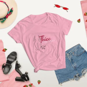 May include: Pink short-sleeved t-shirt with a graphic of a woman's silhouette and a small dog. The text "Thicc Dog Mom" is printed in red on the shirt.
