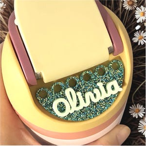 May include: A pastel-colored phone stand with the name "Olivia" in white script. The stand has a glittery teal base and a hinged top. The design incorporates shades of yellow, pink, and purple, creating a playful aesthetic.