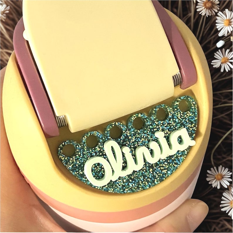 May include: A pastel-colored phone stand with the name "Olivia" in white script. The stand has a glittery teal base and a hinged top. The design incorporates shades of yellow, pink, and purple, creating a playful aesthetic.