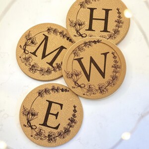 May include: Four round cork coasters with a floral wreath design and individual letters: M, H, W, and E. The letters are dark brown, and the floral wreath is a lighter brown. The coasters are arranged on a white surface.
