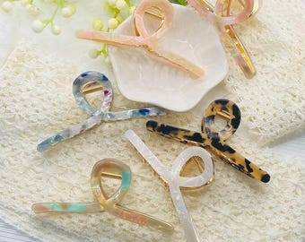 May include: Five hair claw clips with a twisted loop design and gold-toned metal springs. The clips feature various marbled patterns in colours including pink, blue, black, white, and multicoloured pastel tones, arranged on a white lace fabric.