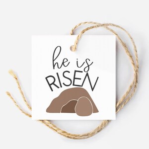 May include: A white gift tag with brown twine. The tag has a brown illustration of a tomb with a large stone rolled away. The text "He is RISEN" is written in black cursive.