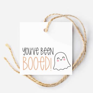 May include: A white square tag with the text "You've Been BOOED!" in black and peach lettering, and a smiling ghost illustration. The tag is tied with a natural twine string, perfect for Halloween.
