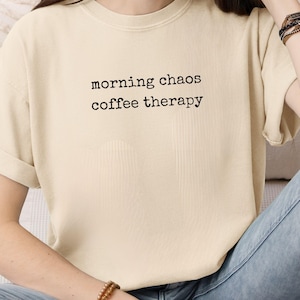Puede incluir: Camiseta de color marfil con el texto "morning chaos coffee therapy" en negro. La camiseta de manga corta está hecha de un material suave. La modelo lleva vaqueros azules y pulseras.