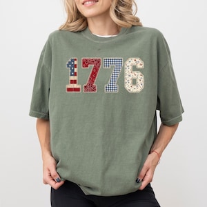 Puede incluir: Una camiseta verde musgo con el número "1776" en un diseño de patchwork. El "1" tiene un patrón de bandera americana, el "77" un patrón de cachemira y el "6" un patrón floral. La camiseta es de corte holgado.