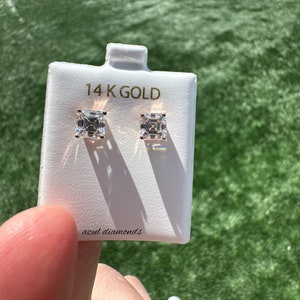 Op de afbeelding: Een paar vierkant geslepen diamant-oorbellen op een witte displaykaart. De kaart leest "14K GOLD" en "azul diamonds". De oorbellen zijn gezet in goud en worden gepresenteerd tegen een groene achtergrond.