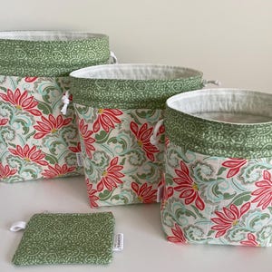 Puede incluir: Conjunto de cuatro recipientes de almacenamiento de tela y una pequeña bolsa. Los recipientes presentan un estampado floral con flores rojas, hojas verdes y detalles azules sobre un fondo crema. Los bordes son verdes con un patrón decorativo.