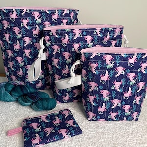 May include: A set of four navy blue fabric bags and a small pouch, all featuring a repeating pattern of pink axolotls and teal plants. The bags vary in size and have pink lining, white drawstrings, and handles. A skein of teal yarn is also present.