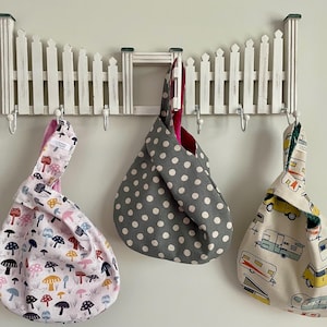 May include: Three fabric knot bags hanging on a white picket fence-style wall rack. The bags feature different patterns: mushrooms, gray polka dots, and camper vans. The bags are in various colors, including pink, gray, and cream.