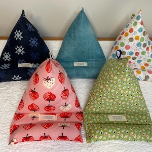 May include: Five triangular fabric book holders. Designs include navy blue with white stars, light blue quilted, pink with strawberries, green floral, and white with yarn balls. Each has a small tag.