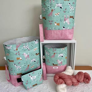 May include: Three fabric storage bags and a small zippered pouch in mint green with a multi-colored dog and flower print. Each bag features a pink textured base, a wrist strap, and a black and white polka dot interior lining. A label reads Cooper's Corner.