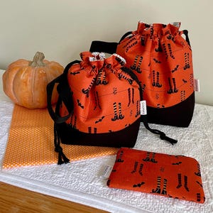 May include: An orange pumpkin sits next to a set of Halloween-themed drawstring bags and a small pouch. The bags and pouch feature a black witch legs and bat pattern on an orange background with black bottoms. The bags have black drawstrings.