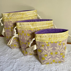 May include: Three drawstring bags in varying sizes. The bags feature a floral pattern in lavender, cream, and yellow. The top band is yellow and white checkered, and the interior is purple. Each bag has a cream-colored handle and a drawstring closure.