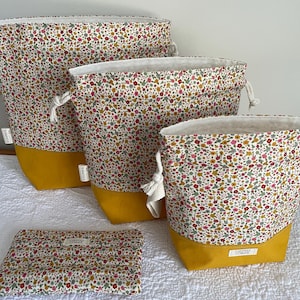 Puede incluir: Juego de cuatro bolsas de almacenamiento con estampado floral y una bolsa a juego. Las bolsas tienen un interior blanco, cierre de cordón y base amarillo mostaza. El estampado floral incluye acentos rojos, rosas y verdes sobre fondo crema.