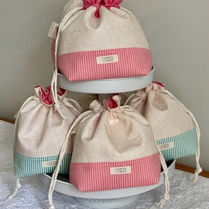 May include: A tiered display of drawstring bags. The bags have a cream-colored background with a subtle swirl pattern and a pink or teal striped bottom. Each bag has a pink ruffled top and a white drawstring. The text "COOPER'S CORNER" is visible on a tag on each bag.