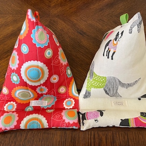 May include: Two triangular fabric book holders. One is red with a floral pattern in orange, blue, and white. The other is off-white with dog illustrations wearing sweaters. Both have a tag that reads "COOPER'S CORNER".