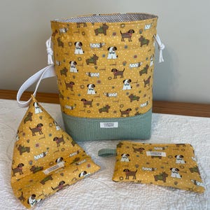 Può includere: Un set di articoli in tessuto a tema cani, tra cui un contenitore alto con chiusura a coulisse, un porta libri triangolare e una piccola pochette con cerniera. Gli articoli presentano uno sfondo giallo con illustrazioni di cani ripetute e un motivo a spina di pesce verde.
