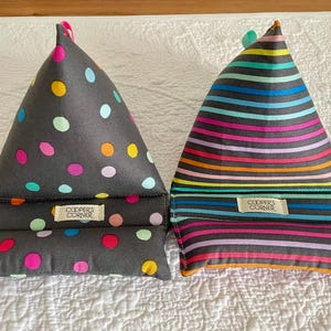 Puede incluir: Dos soportes triangulares de tela para libros. Uno es gris oscuro con lunares de colores, el otro tiene rayas horizontales de arcoíris. Ambos tienen una pequeña etiqueta que dice "COOPER'S CORNER".