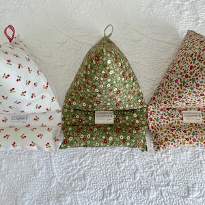 May include: Three fabric book holders with floral and cherry patterns. The holders are triangular, with a loop at the top and a rectangular base. Each has a tag that reads "COOPER'S". The patterns include cherries on white, small flowers on green, and small flowers on a cream background.