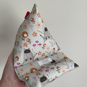 Peut inclure: Un support triangulaire en tissu pour tablette ou téléphone avec un fond bleu clair et un motif répétitif de chats siamois, de fleurs orange et de fleurs roses. Une étiquette blanche avec le texte COOPERS CORNER est cousue sur le devant.
