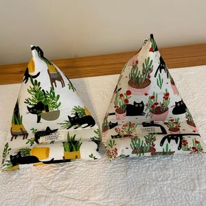 Op de afbeelding: Twee driehoekige stoffen tablethouders met een witte achtergrond, voorzien van illustraties van zwarte katten en kamerplanten. Elke houder heeft een klein wit label met de tekst COOPER'S CORNER. De patronen bevatten diverse groene planten en rode bloemen.