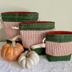 Op de afbeelding: Set van drie stoffen opbergmanden met een pompoenpatroon, groene rand en crèmekleurige handvatten. Twee pompoenen, één oranje en één wit, zijn voor de manden geplaatst. De manden hebben een rode binnenkant.