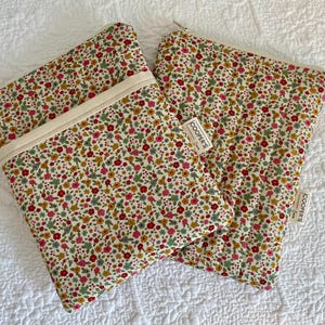 Könnte beinhalten: Zwei Reißverschlusstaschen mit floralem Druck und cremefarbenem Besatz und Reißverschluss. Die Taschen weisen ein Muster aus kleinen roten, rosa, gelben und grünen Blumen auf cremefarbenem Hintergrund auf. Jede Tasche hat ein kleines Etikett mit der Aufschrift "COOPER'S".