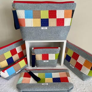 May include: A collection of five gray zippered pouches in varying sizes, each featuring a patchwork panel of colorful patterned squares. Each pouch includes a red zipper closure and a dark blue wrist strap attached to a silver metal ring.